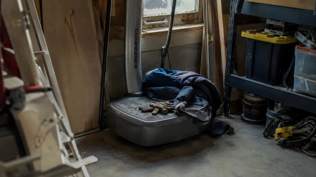 A vibration plate sitting unused in the corner of a garage collecting dust with a coat draped over it